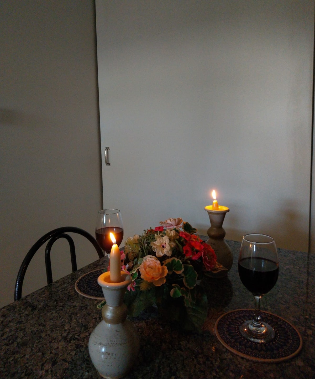 A dining table is set with two glasses of red wine beside a floral arrangement featuring soft-colored blooms. Candles in beige holders provide a warm glow, enhancing the tranquil setting. A smooth granite surface reflects the gentle light, creating a cozy dining atmosphere.