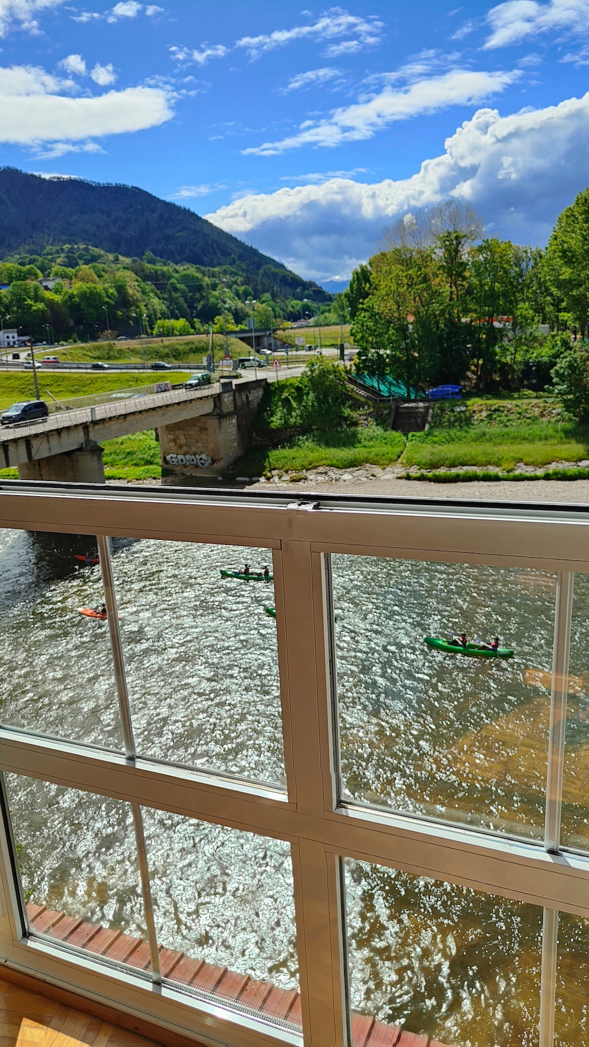 A view of the river Sella is captured from a large window, showcasing the flowing water and canoeists paddling nearby. Lush green hills and a distant bridge are visible, framed by a bright blue sky with clouds, creating a serene outdoor scene.