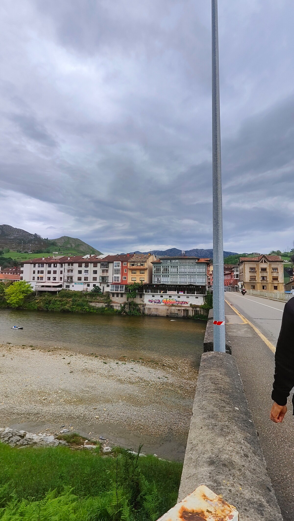 The image captures a view from a bridge overlooking the Sella River. Colorful buildings line the riverbank, showcasing a mix of architectural styles. Lush greenery surrounds the water, while a cloudy sky looms above, reflecting the tranquil nature of the setting.