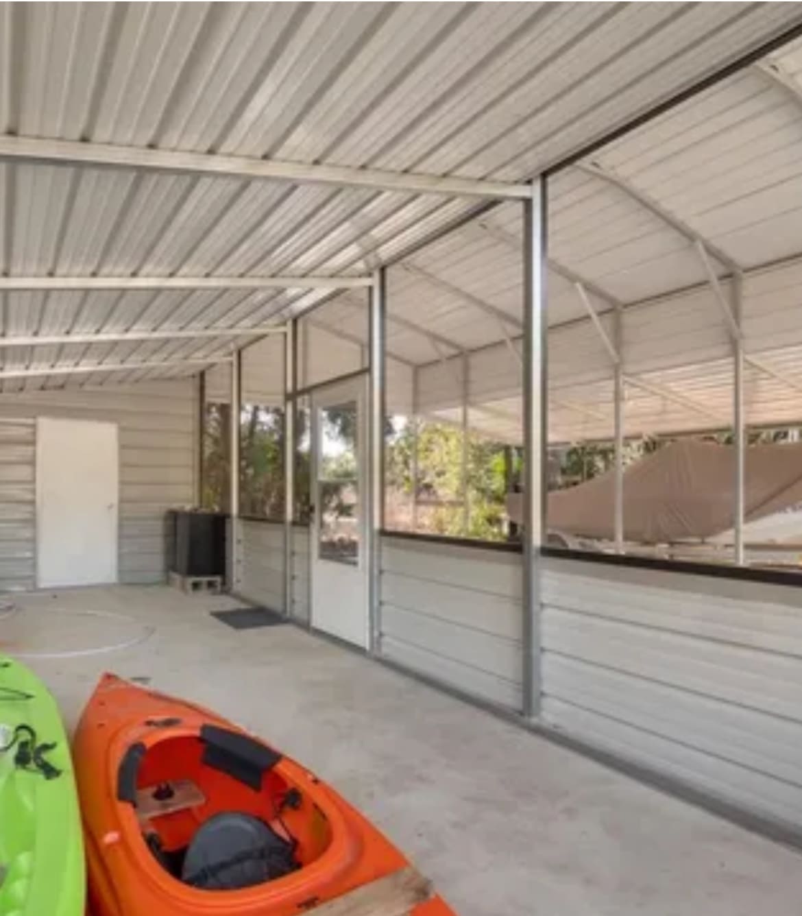 A screened lanai is presented, featuring a concrete floor and metal framework. Two kayaks in green and orange are positioned on the left, while a white door leads outside on the right. Natural light filters in through the screens, showcasing the surrounding greenery.