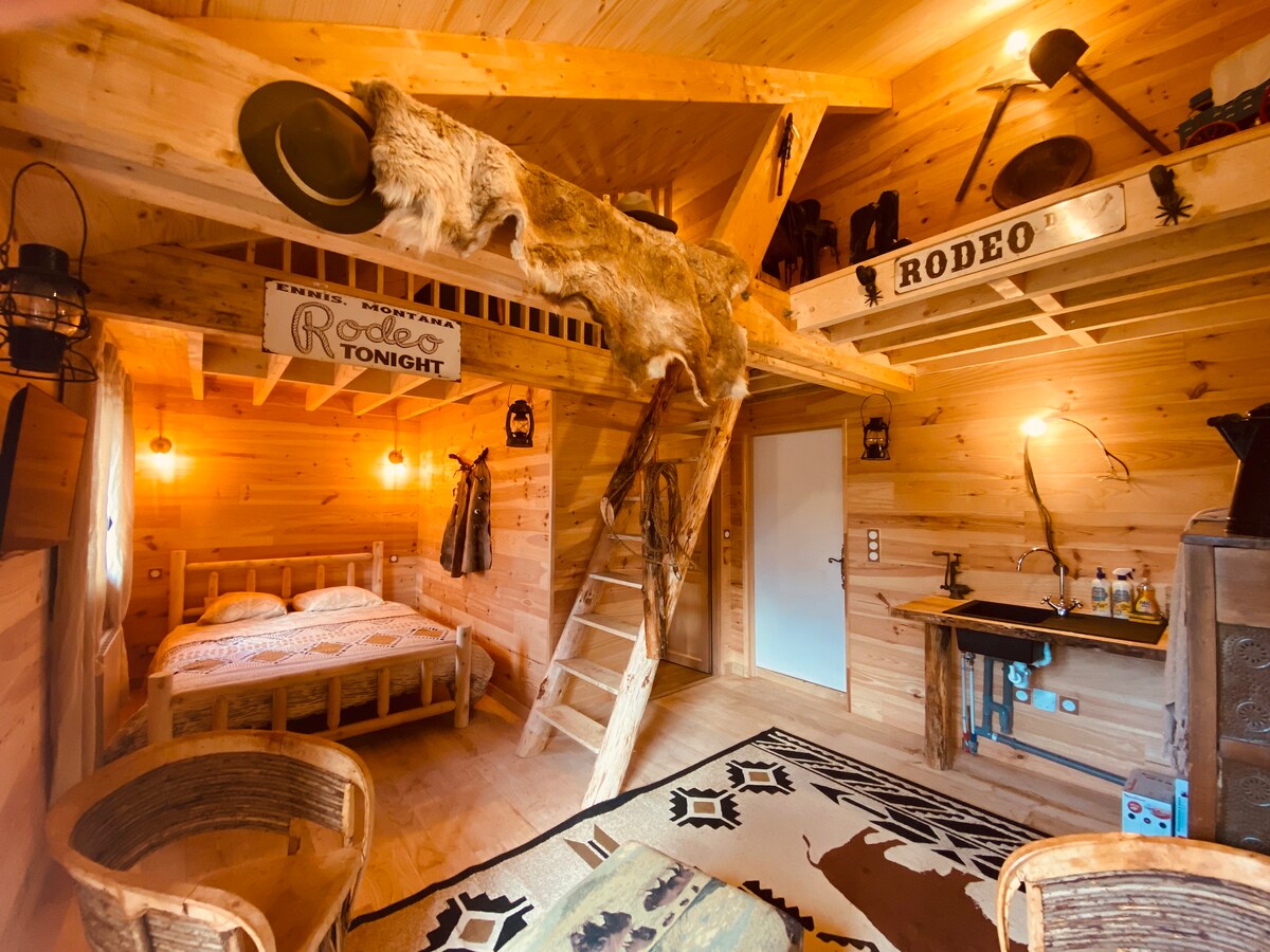 A rustic interior features wooden walls and ceiling, illuminated by warm lighting. A spacious bed is situated next to a wooden ladder leading to an upper sleeping area. Various cowboy-themed decor items, including a mounted hide and signs, enhance the charming ambiance.