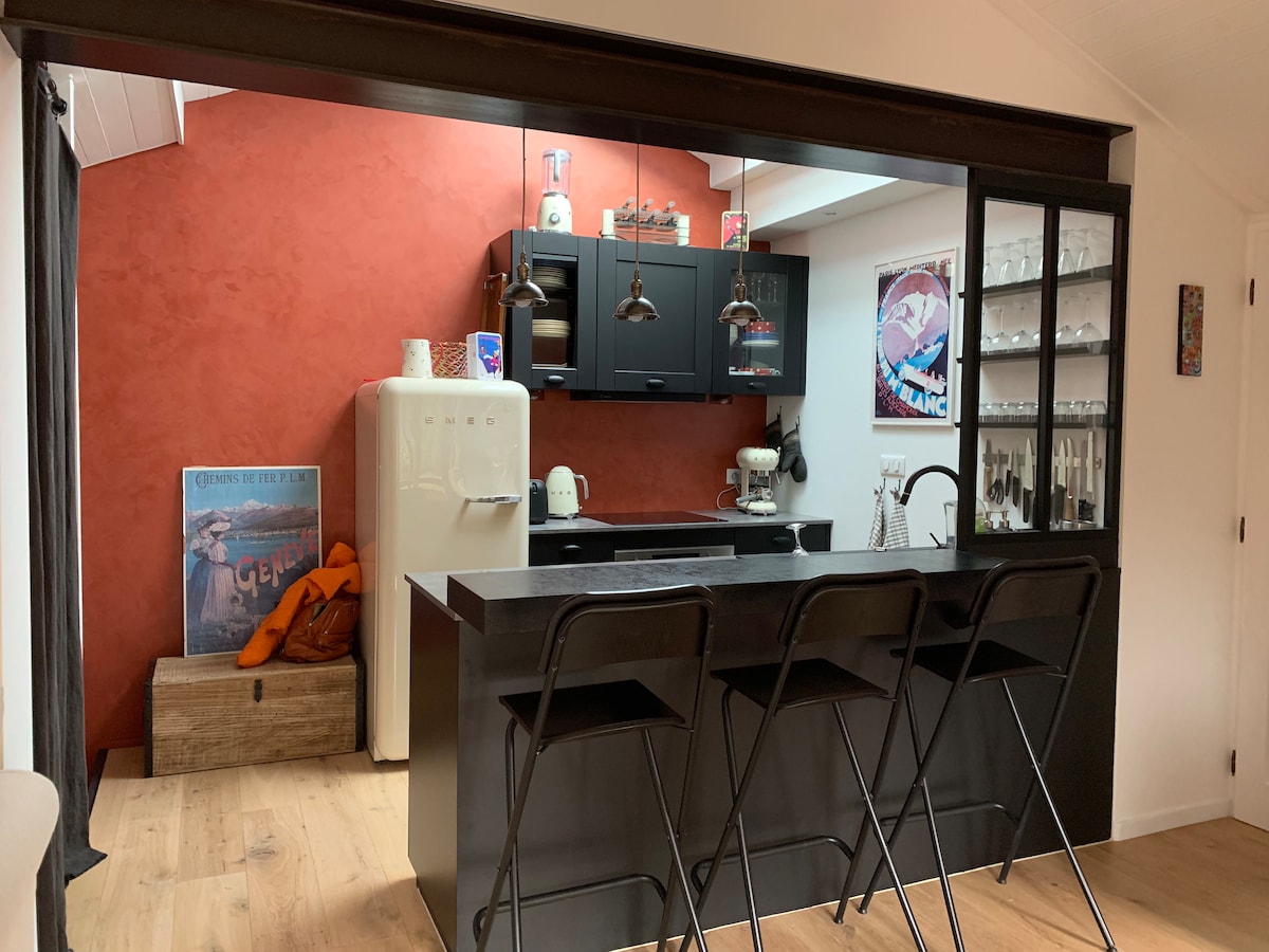An open kitchen area is highlighted by a red accent wall and black cabinetry. A central island with three black bar stools is visible. Shelves containing glassware are mounted on the wall, while vintage appliances add charm to the space.