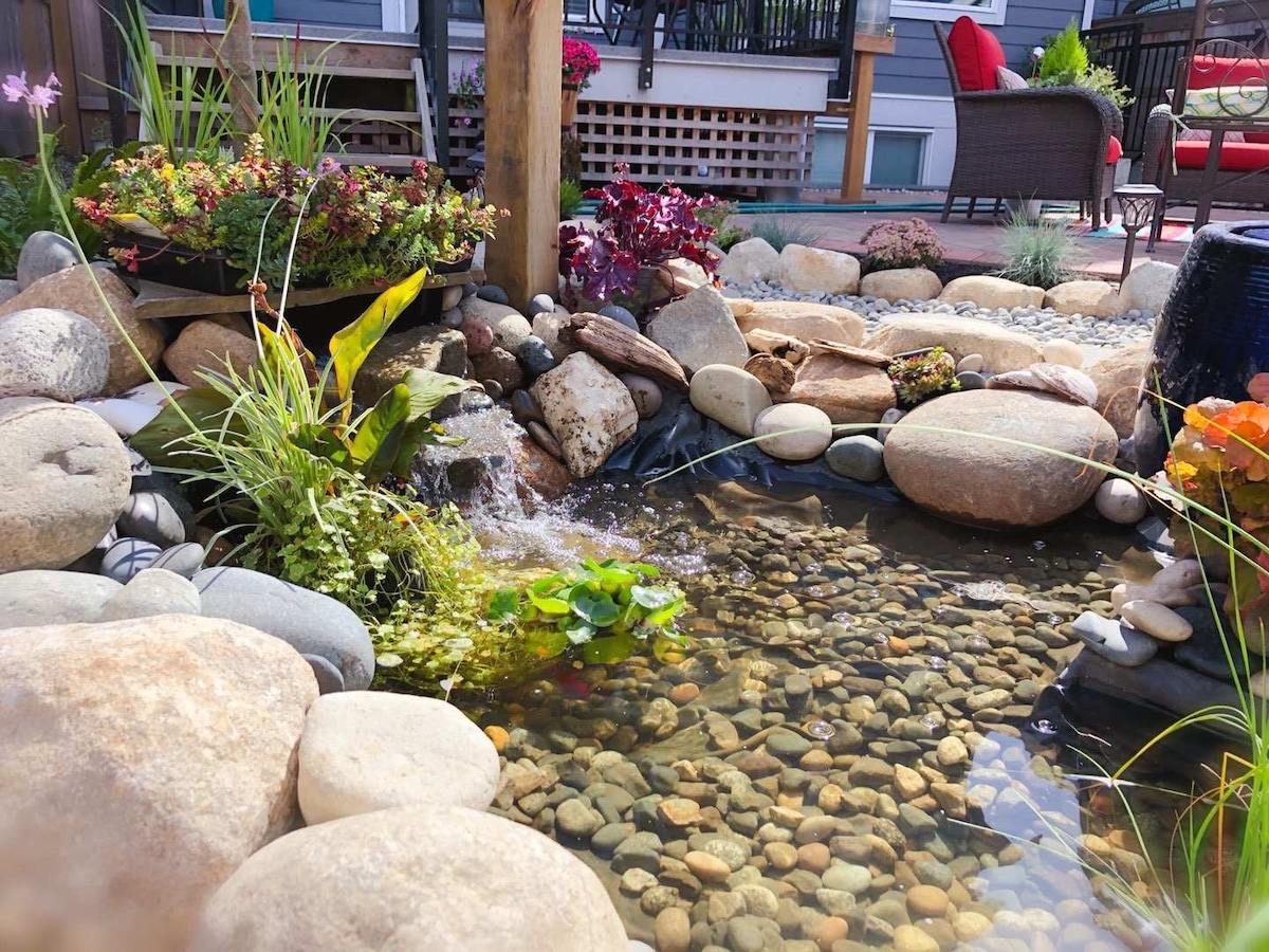 A tranquil garden area is featured, showcasing a small pond filled with smooth stones and water plants. Water gently cascades over rocks, surrounded by lush greenery and colorful flowers. The inviting landscape is complemented by a seating area visible in the background.