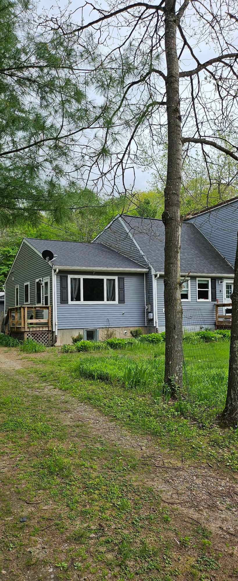 A charming exterior view of a single-story house is presented, surrounded by greenery and trees. The home features a light-colored siding, a sloped roof, and a small porch area. A gravel path leads up to the entrance, enhancing the natural setting.