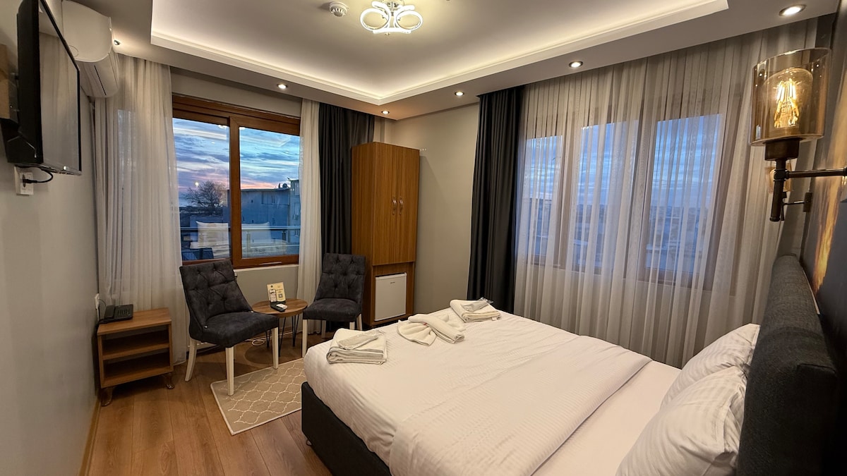 A modern bedroom features a spacious bed with neatly arranged linens. Two cozy chairs are positioned beside a wooden cabinet. Large windows allow natural light to fill the room, showcasing a pleasant view outside. Soft lighting adds to the comfortable ambiance.