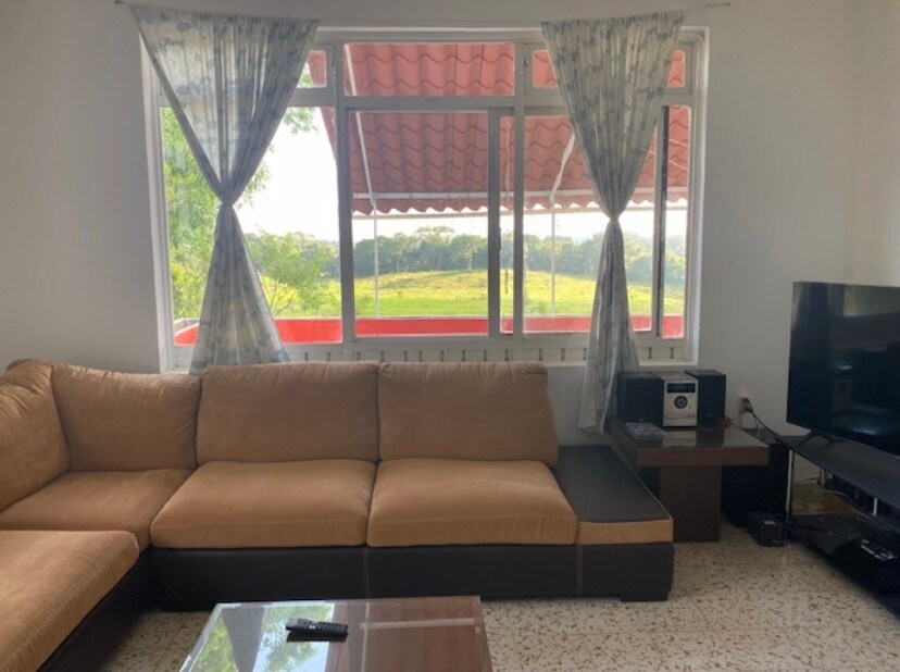 A spacious living area features a large, L-shaped sofa that invites relaxation. Natural light enters through wide windows adorned with sheer curtains, providing views of the greenery outside. A coffee table rests on the tiled floor, and a television is positioned on a nearby stand.