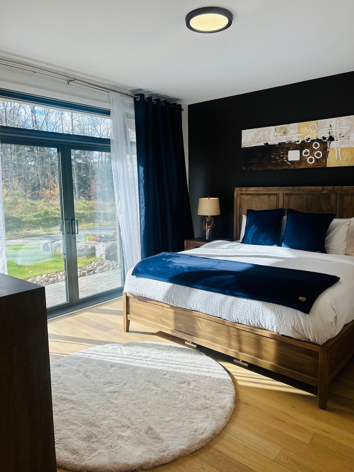 A master bedroom features a king-size bed with plush bedding and accent pillows in deep blue. Natural light floods in through floor-to-ceiling glass doors, leading to a serene outdoor view. A soft area rug is placed beneath the bed, and dark curtains frame the windows.