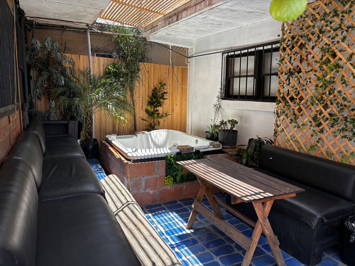 An outdoor patio area features a hot tub surrounded by potted plants and wood paneling. Comfortable seating is provided by black leather sofas, and a wooden table is positioned in the center, contributing to a serene and relaxing environment.