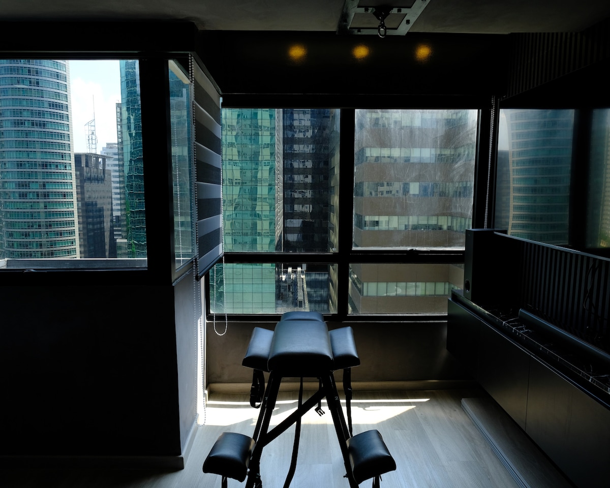 A bright space is showcased with large windows offering views of surrounding skyscrapers. A massage table is positioned centrally, while contemporary cabinetry complements the modern decor. Natural light enhances the room's spacious feel, creating a calming atmosphere for relaxation.