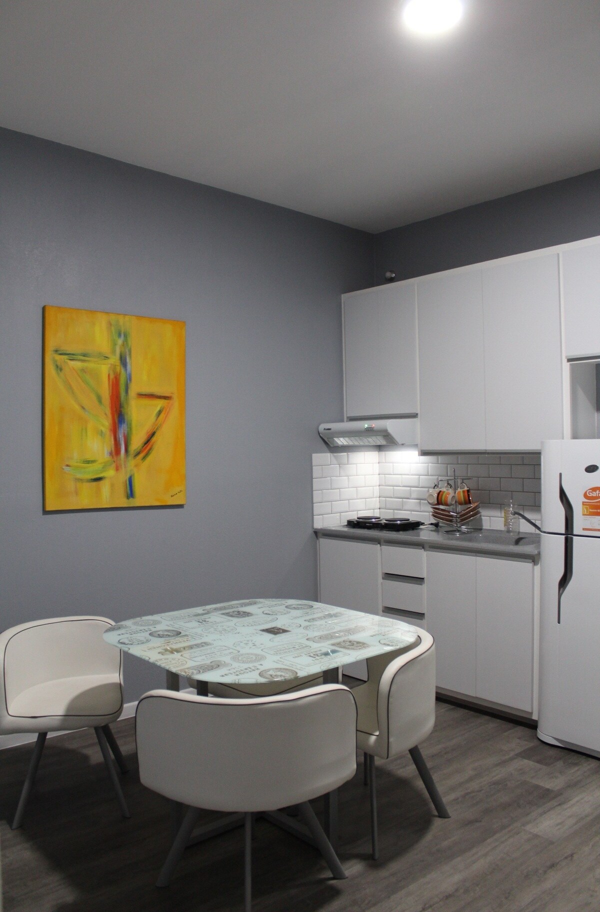 A compact kitchen area features a white table surrounded by four chairs. A colorful painting hangs on the gray wall beside modern cabinetry and a cooking area with a hood. Natural light reflects off the sleek surfaces, creating a bright and functional space.