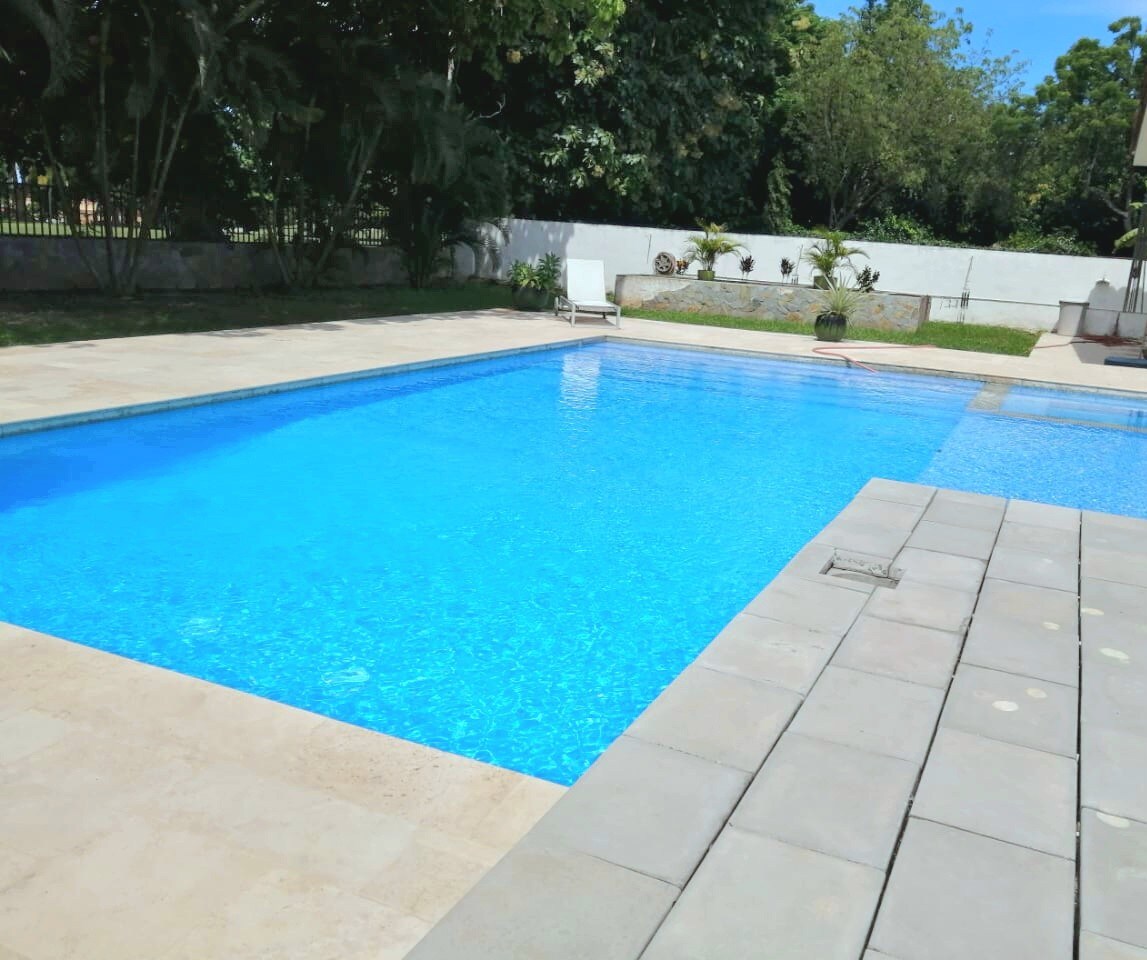 A sparkling pool is surrounded by a spacious patio, featuring a sun lounge chair positioned for relaxation. Lush greenery and trees provide a natural backdrop, creating a serene outdoor environment. The pool area benefits from ample sunlight, enhancing the inviting atmosphere.
