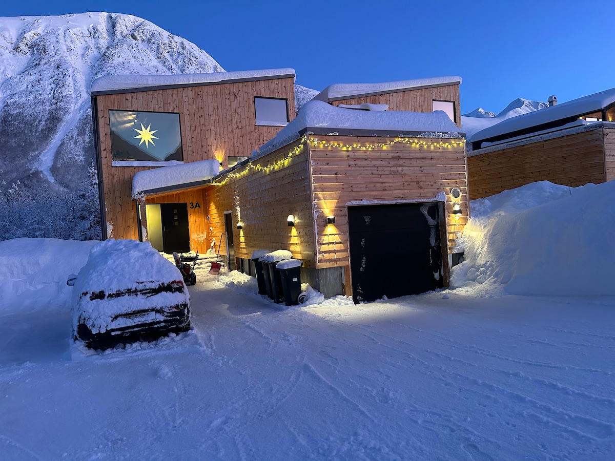 A contemporary wooden house is gently illuminated by exterior lights, surrounded by a snowy landscape. The entrance features a large star decoration in the window, with snow covering the ground and nearby vehicles. A garage door is visible, adding to the home's modern design.