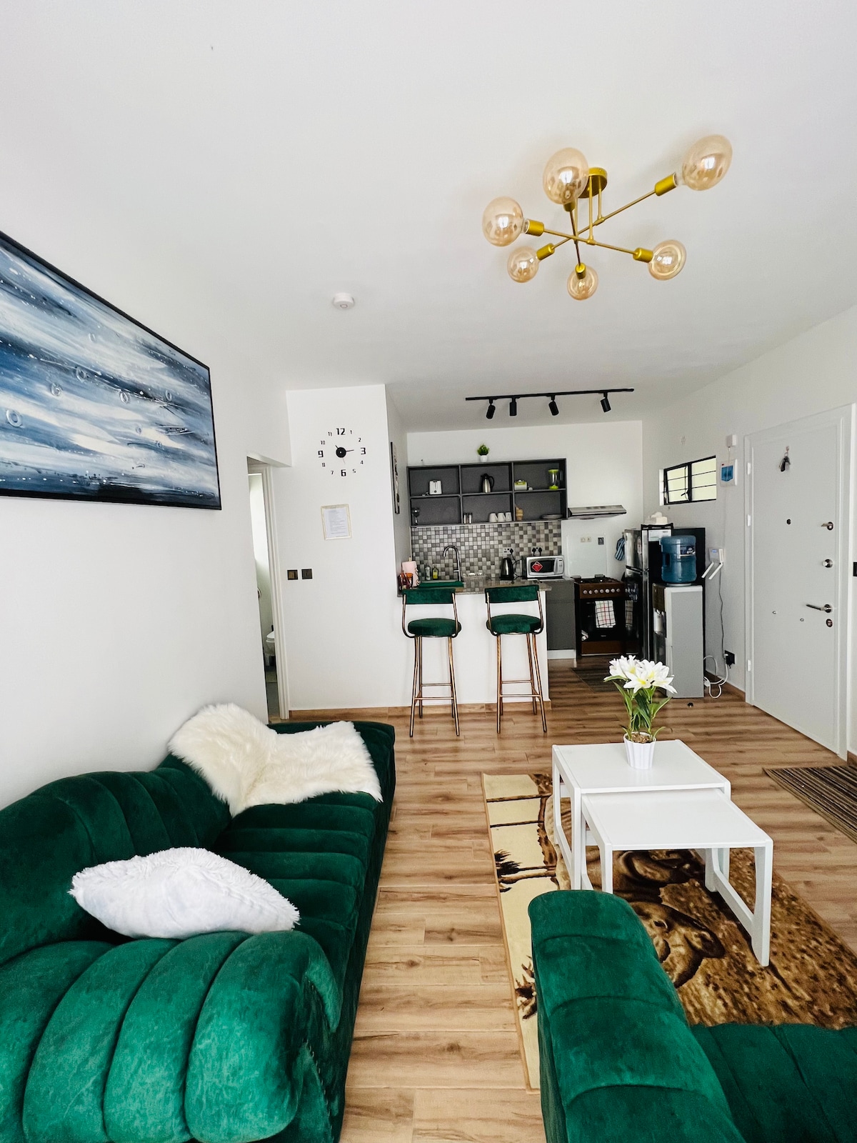 A modern living area features a vibrant green sofa paired with a plush white throw. A white coffee table sits on a decorative rug. The kitchen area is visible in the background, showcasing a sleek design with bar stools and contemporary appliances.