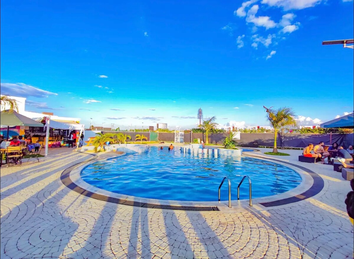 A large, circular pool is surrounded by patterned stone decking, featuring several lounge areas with umbrellas. Palm trees provide shade, and the skyline is visible in the background under a clear blue sky. Sunlight reflects off the water, adding to the calm environment.