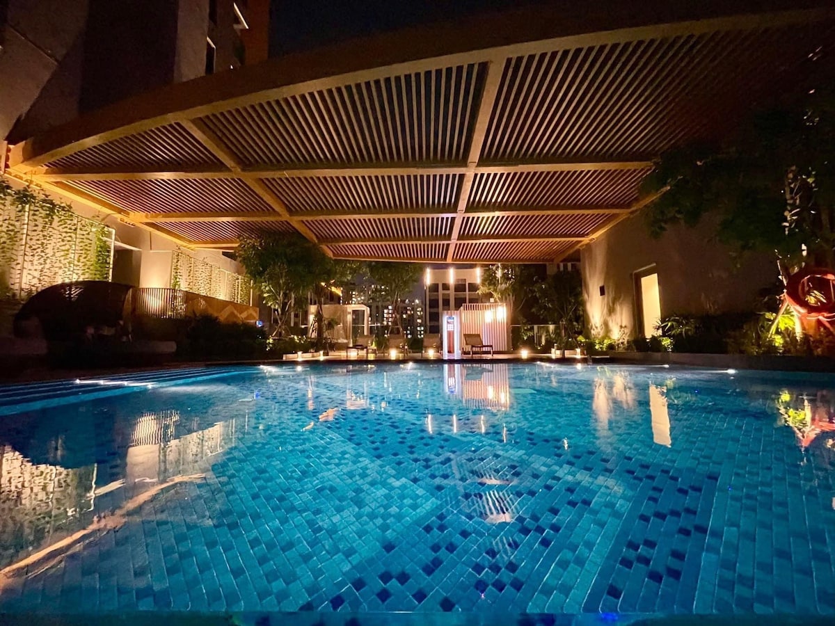 A tranquil swimming pool is illuminated at night, surrounded by green plants and decorative lighting. The water's surface reflects the intricate tile patterns and the architectural features of the overhead structure. Soft ambient light enhances the serene atmosphere of the space.