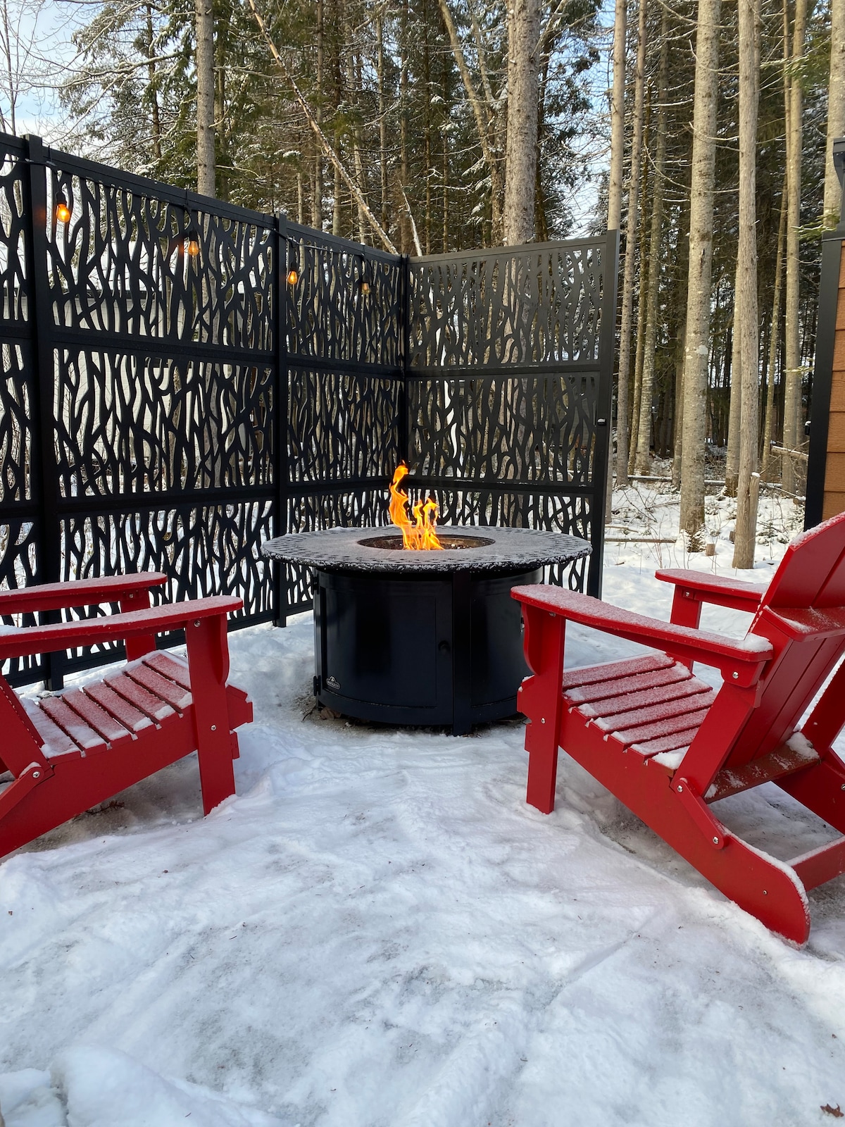 An outdoor propane fire pit is surrounded by two bright red chairs. The area is enclosed by a decorative black screen, offering a cozy space. Snow blankets the ground, and tall trees are visible in the background, creating a serene winter setting.