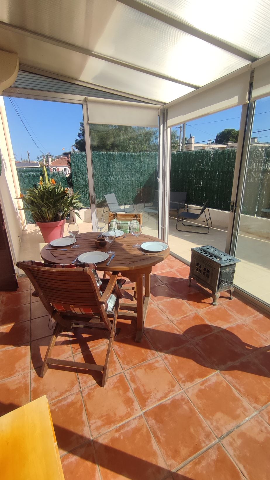 A sunlit dining area features a round wooden table set for four, surrounded by folding chairs. A potted plant is displayed nearby, and large glass doors open to a private outdoor space. Bright tiles create a warm ambiance with natural light streaming in.