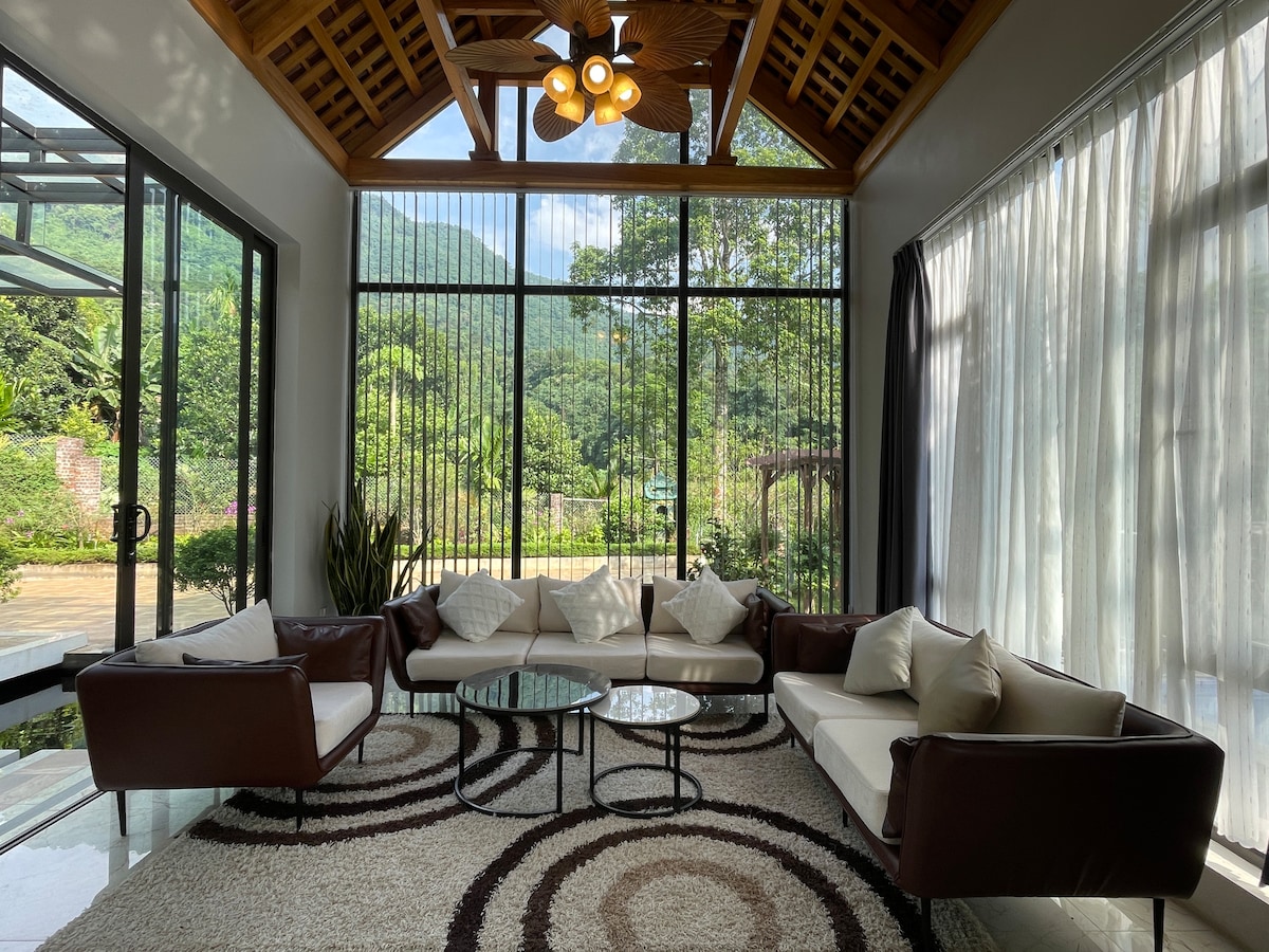 A modern living area features large glass windows framing a scenic view of the surrounding greenery. Two brown sofas with cream cushions are arranged around a circular glass table, set atop a plush area rug with a circular pattern. Natural light floods the space, highlighting its open layout.