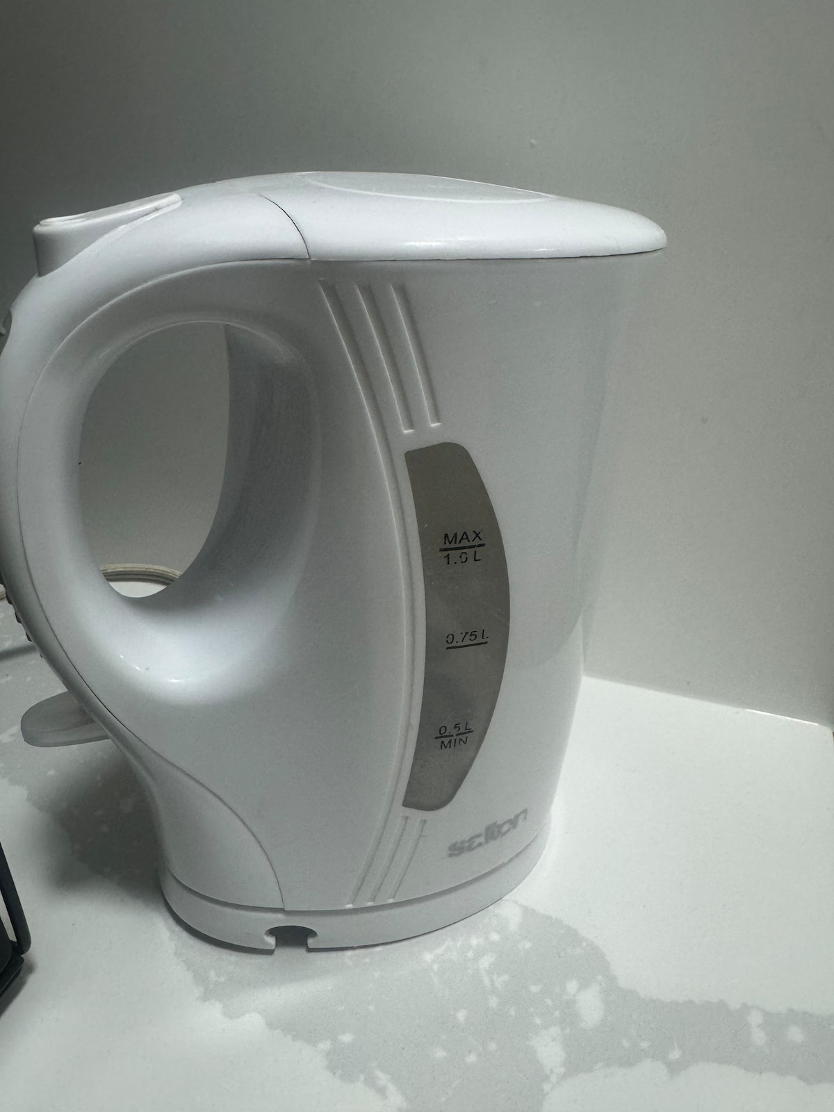 An electric kettle is positioned on a light-colored countertop. The kettle features a sleek design with a clear water level indicator, marked with measurements up to 1.9 liters. Its white exterior complements the minimalist kitchen decor.