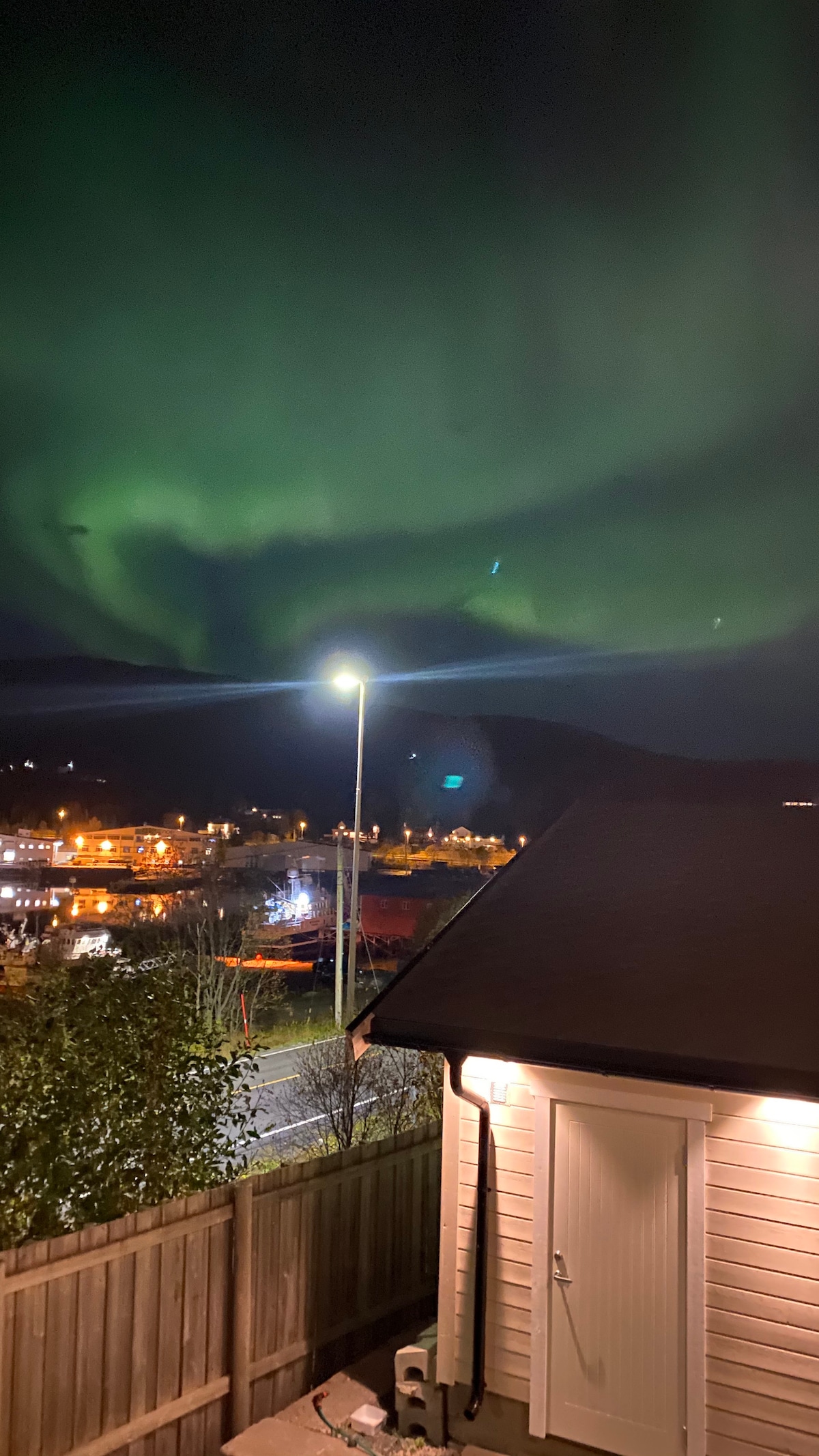 The night sky is illuminated by vibrant green auroras, cascading above the landscape. Below, soft glow from nearby lights creates a warm contrast against the natural display, while a corner of a building is visible to the right. The serene setting suggests a peaceful ambiance.