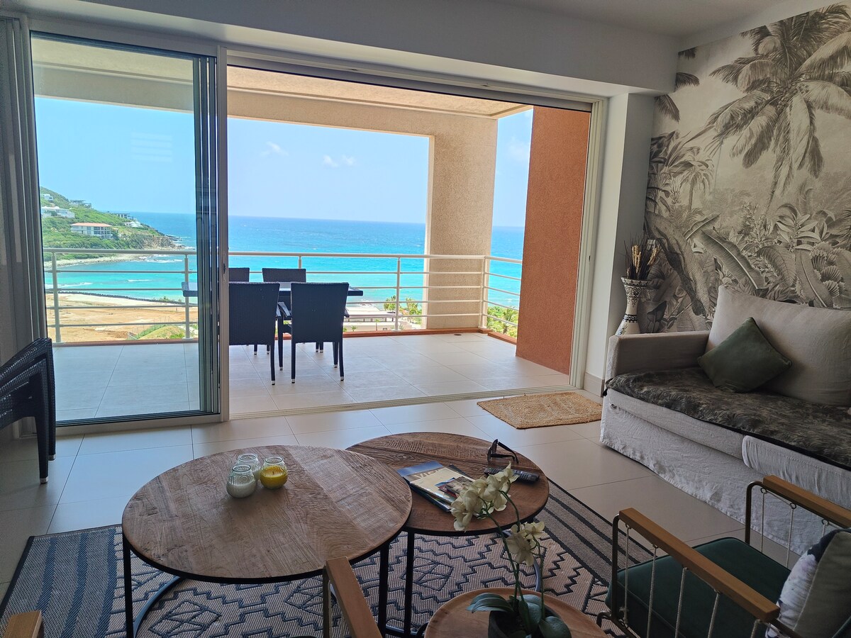 A spacious living area features large sliding glass doors that open to a balcony with ocean views. Comfortable seating is arranged around a circular table, while decorative plants and a neutral color palette enhance the serene vibe. Natural light floods the room, creating a bright atmosphere.