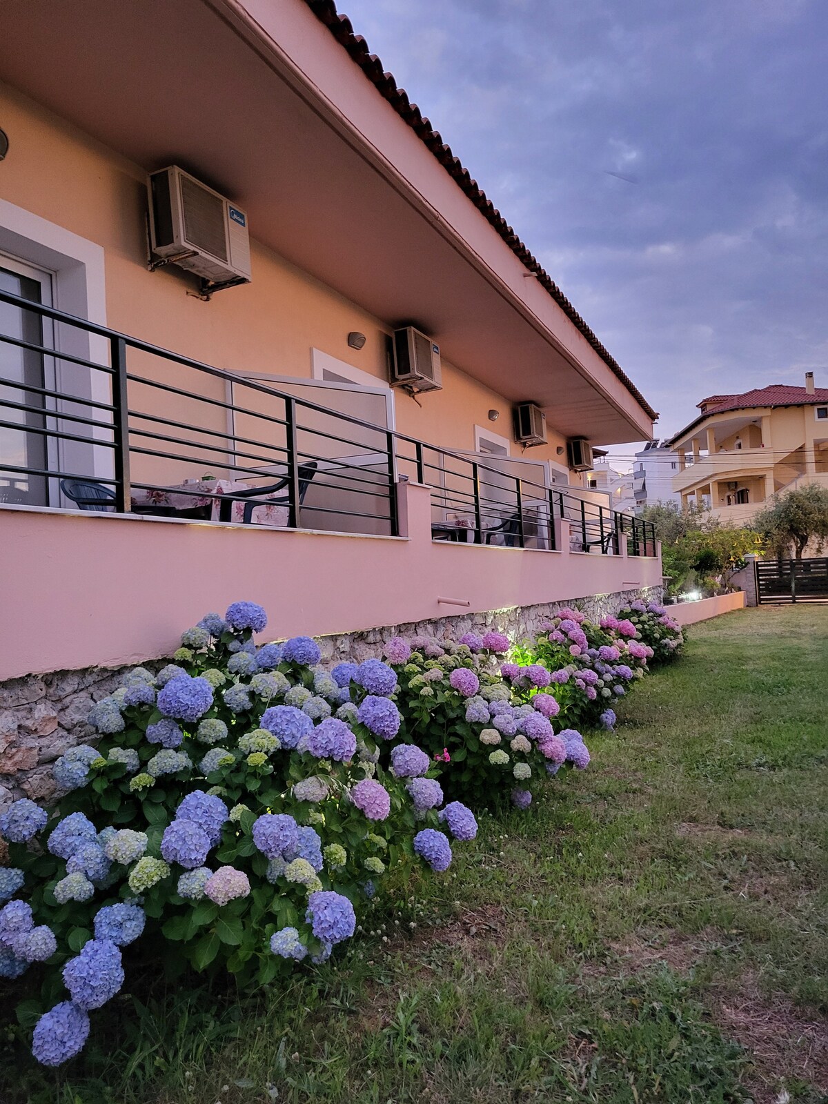 A garden area is showcased, with vibrant hydrangea flowers in shades of blue and purple lining a stone wall. The building's exterior is visible, featuring air conditioning units and a balcony area that overlooks the garden.