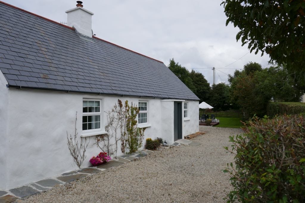 A charming irish cottage presents a whitewashed exterior and slate roof, surrounded by a gravel pathway. Green hedges and blooming plants add contrast to the facade, while a glimpse of the garden and outdoor seating area is visible in the background.