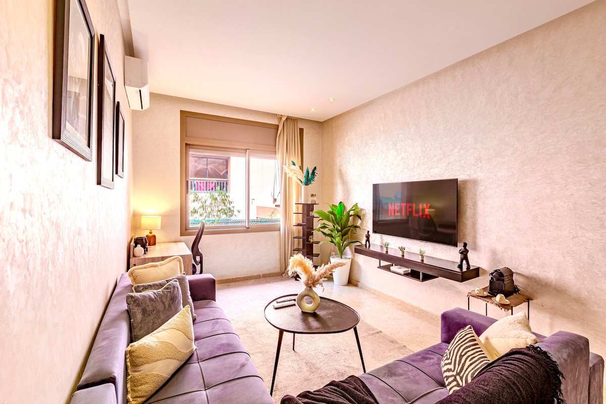 A comfortable living area features a large TV mounted on the wall and two purple sofas surrounding a circular coffee table. Natural light enters through the window, illuminating a potted plant and an elegant decorative piece on the side. The walls are adorned with framed artwork.