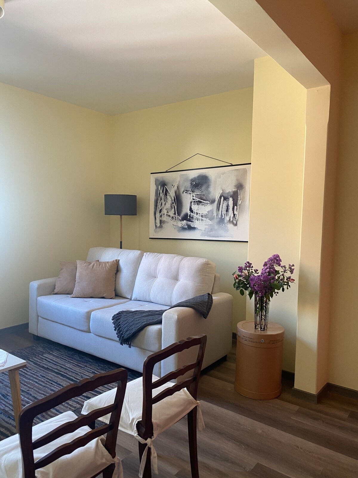 A light-filled living area features a comfortable sofa adorned with decorative pillows. A piece of artwork hangs on the wall above, complemented by a floor lamp. A vase with fresh flowers adds a touch of color, while a woven rug grounds the space.