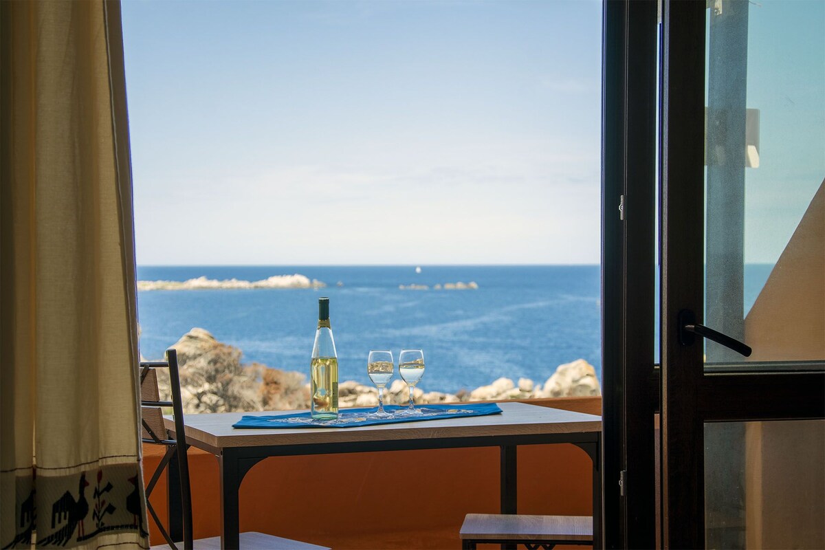 A serene dining space is presented, featuring a table set for two with a bottle of wine and glasses. Through the open glass doors, views of the tranquil sea and distant rocks are visible, inviting relaxation and enjoyment of the coastal scenery.