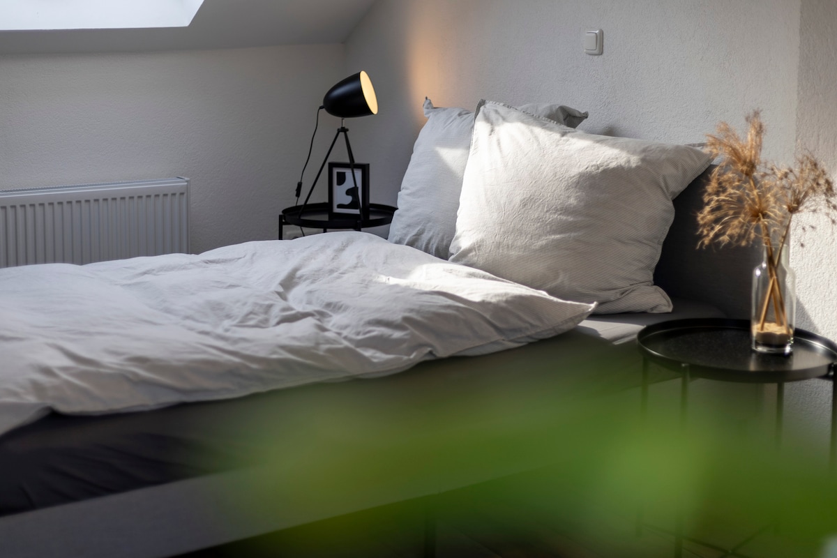 A cozy sleeping area features a spacious bed adorned with soft, neutral bedding. A bedside table holds a stylish lamp and a small decorative vase, while natural light filters through a nearby window. Greenery adds a touch of warmth to the space.
