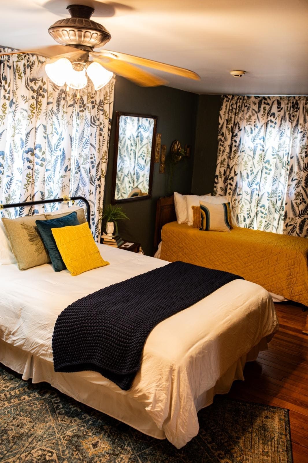 A cozy sleeping area features a spacious bed topped with a white comforter and a navy throw blanket. A bright accent pillow is displayed on the bed, while a second bed is positioned nearby. The room is illuminated by ceiling fans, with floral-patterned curtains framing the windows.