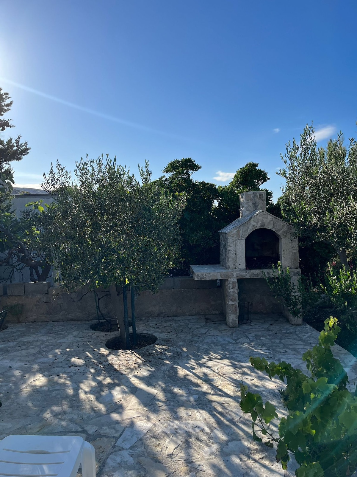 A sunlit outdoor area showcases a stone barbecue grill surrounded by lush greenery. Olive trees provide shade, and sunlight filters through the branches, casting gentle shadows on the stone-paved ground, creating a peaceful atmosphere for outdoor gatherings.