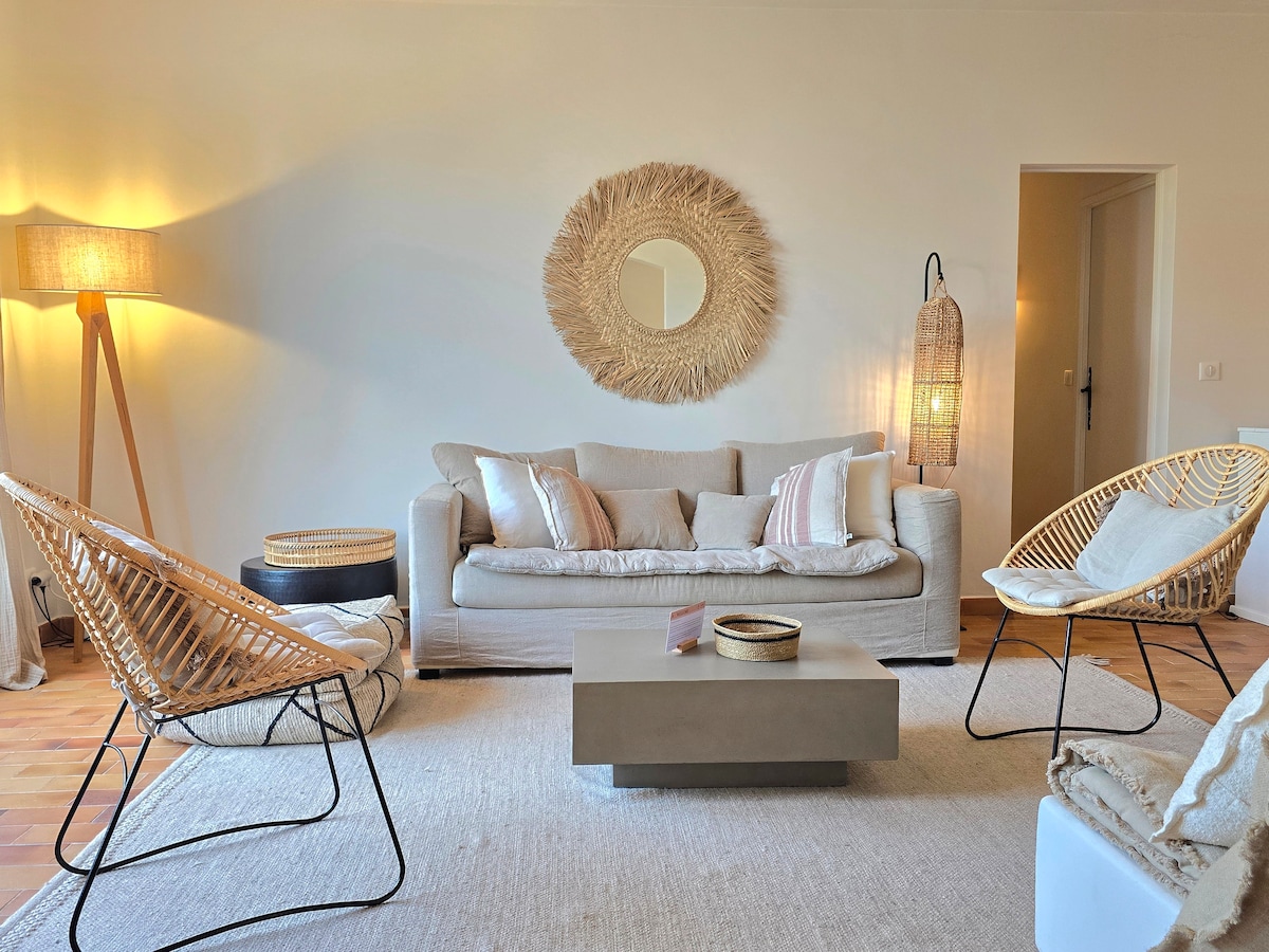 The living area features a soft sofa adorned with pastel pillows, complemented by two wicker armchairs. A round mirror enhances the wall, while a central coffee table provides a functional touch. Warm light is emitted by a standing lamp, creating a cozy atmosphere.