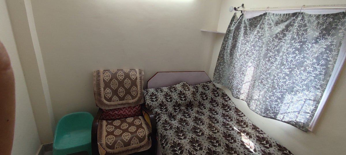 A modest sleeping area features a spacious bed covered with a floral-patterned blanket. A comfortable armchair is positioned nearby. Natural light enters through a window, partially covered by light-patterned curtains, creating a calm and relaxing ambiance.