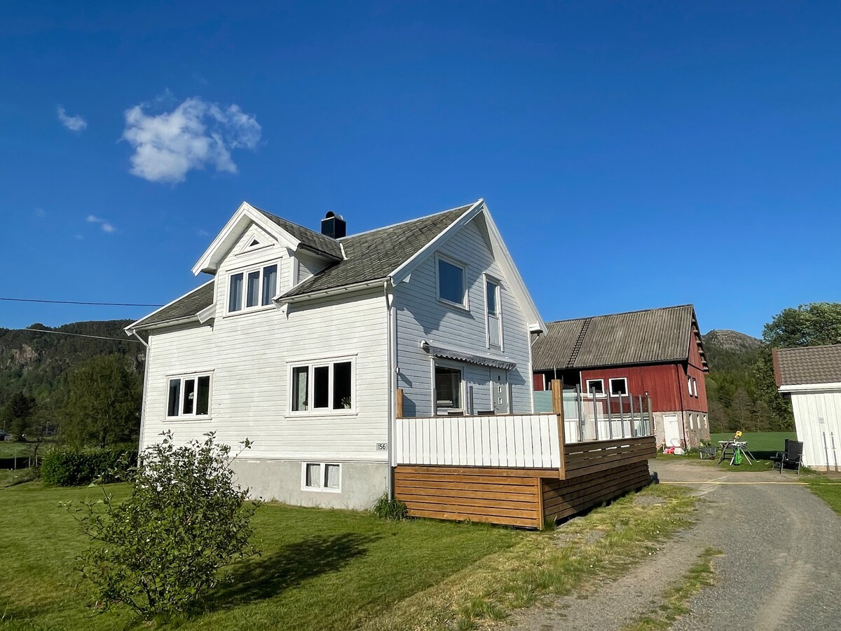 A two-story house is shown with a light exterior, alongside a red barn structure in the background. A wooden deck extends from the house, and a gravel path leads to the entrance. Green grass surrounds the property, complemented by a clear blue sky.