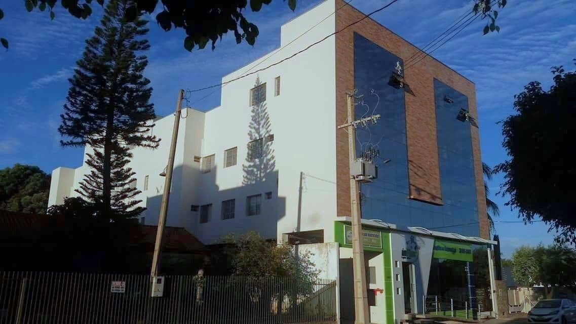 The exterior of a modern flat building is shown, with large windows reflecting the sky. A green entrance can be seen at the base, surrounded by landscaping and trees. Power poles and lines are present in the foreground, indicating a well-connected location.