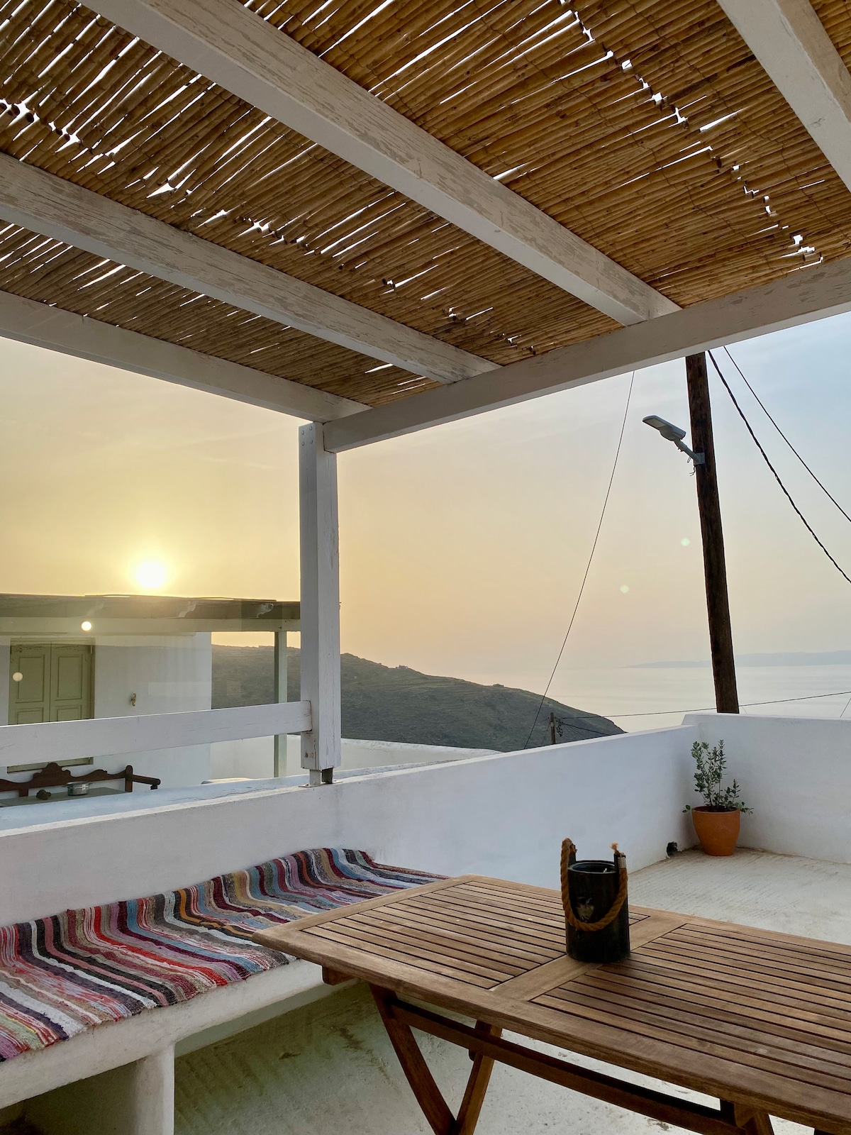 An outdoor terrace is visible, featuring a wooden table surrounded by a multi-colored woven seating area. The sun sets in the distance, casting warm hues over the landscape. A small potted plant adds a touch of greenery beside the seating. Light is gently diffused through a bamboo canopy overhead.