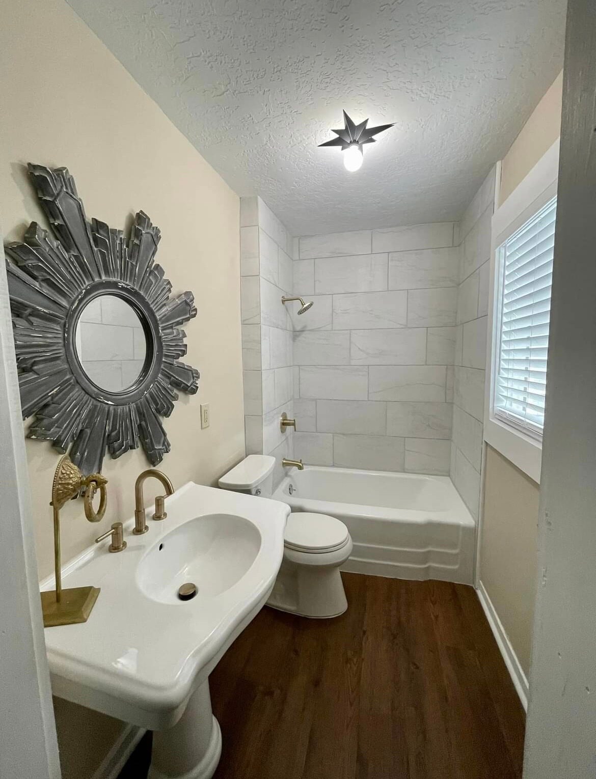 A well-appointed bathroom features white marble tile surrounds with a shower-tub combination. A large decorative mirror with a sunburst design reflects light. Elegant gold fixtures and a white pedestal sink integrate seamlessly with rich brown flooring.