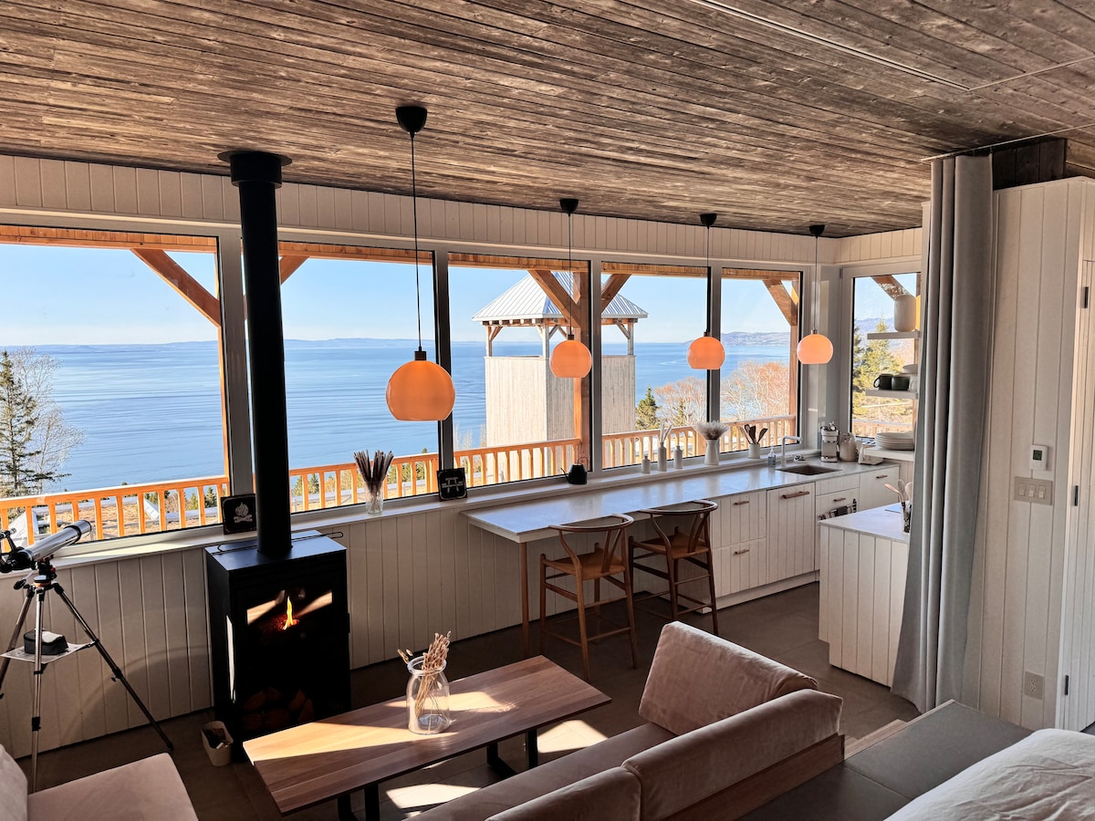 An open and airy kitchen area is showcased, featuring a large window overlooking a serene body of water. Warm pendant lights hang above a wooden table, while a cozy seating area with a soft couch is positioned nearby. A sleek wood stove adds warmth to the space.