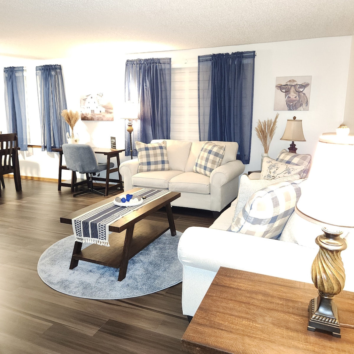 The living area features a comfortable seating arrangement with neutral sofas adorned with plaid accent pillows. A coffee table is centered on a round blue rug, and natural light filters through the window drapes, creating a calm ambiance.