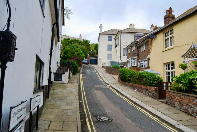 A narrow, upward-sloping street is flanked by quaint houses featuring varied architectural styles. Paved pathways and yellow road markings guide towards the horizon, while greenery lines the sides, adding a natural touch. The scene conveys a peaceful neighborhood atmosphere in a historic town.