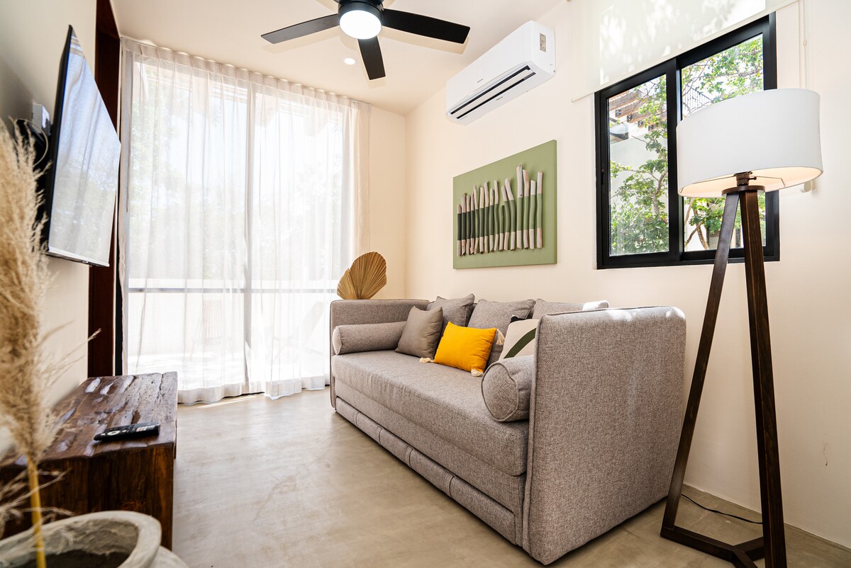 A light-filled living area features a comfortable gray sofa adorned with a bright yellow accent pillow. A wooden coffee table rests in front, complemented by a standing lamp. Air conditioning is provided, while expansive windows offer views of greenery outside.