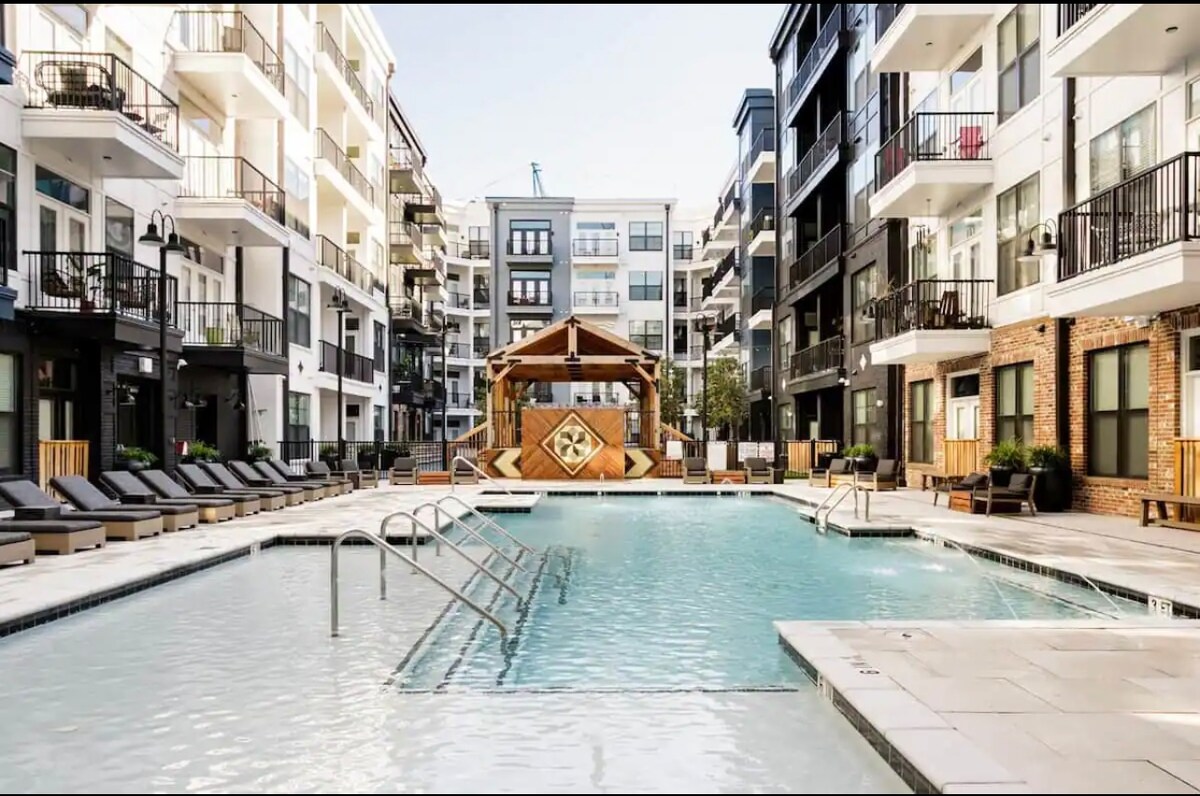 A spacious outdoor pool is framed by modern apartment buildings, featuring lounge chairs arranged around the edge. A wooden gazebo is situated at one end of the pool area, creating a welcoming communal space. Clear skies reflect in the calm water.