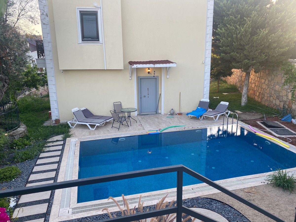 A private pool area features clear blue water, surrounded by lounge chairs and a small table. A pathway lined with stones leads towards the house, which is painted in light yellow and includes a welcoming door. Lush greenery enhances the tranquil setting.