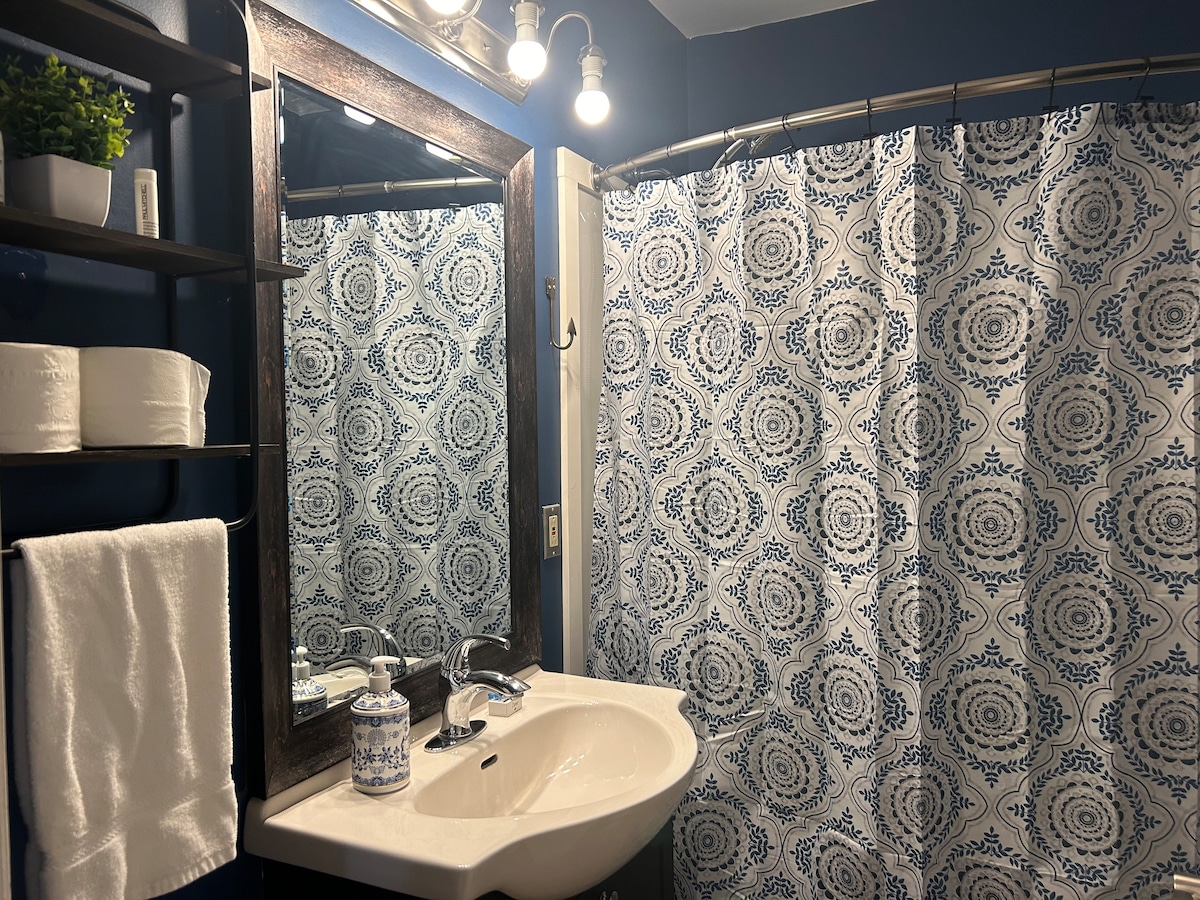 A bathroom is presented with a dark blue color scheme and decorative wallpaper. A white sink is positioned beneath a mirrored cabinet, flanked by neatly arranged towels. A patterned shower curtain adds a splash of color, while shelves display decorative items and toiletries.