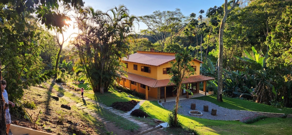 A spacious two-story house featuring a warm yellow exterior is set against a lush green landscape. The surrounding area includes tall trees and manicured grass, with a pathway leading to the entrance. A seating area with stone seating circles the firepit, inviting outdoor gatherings.