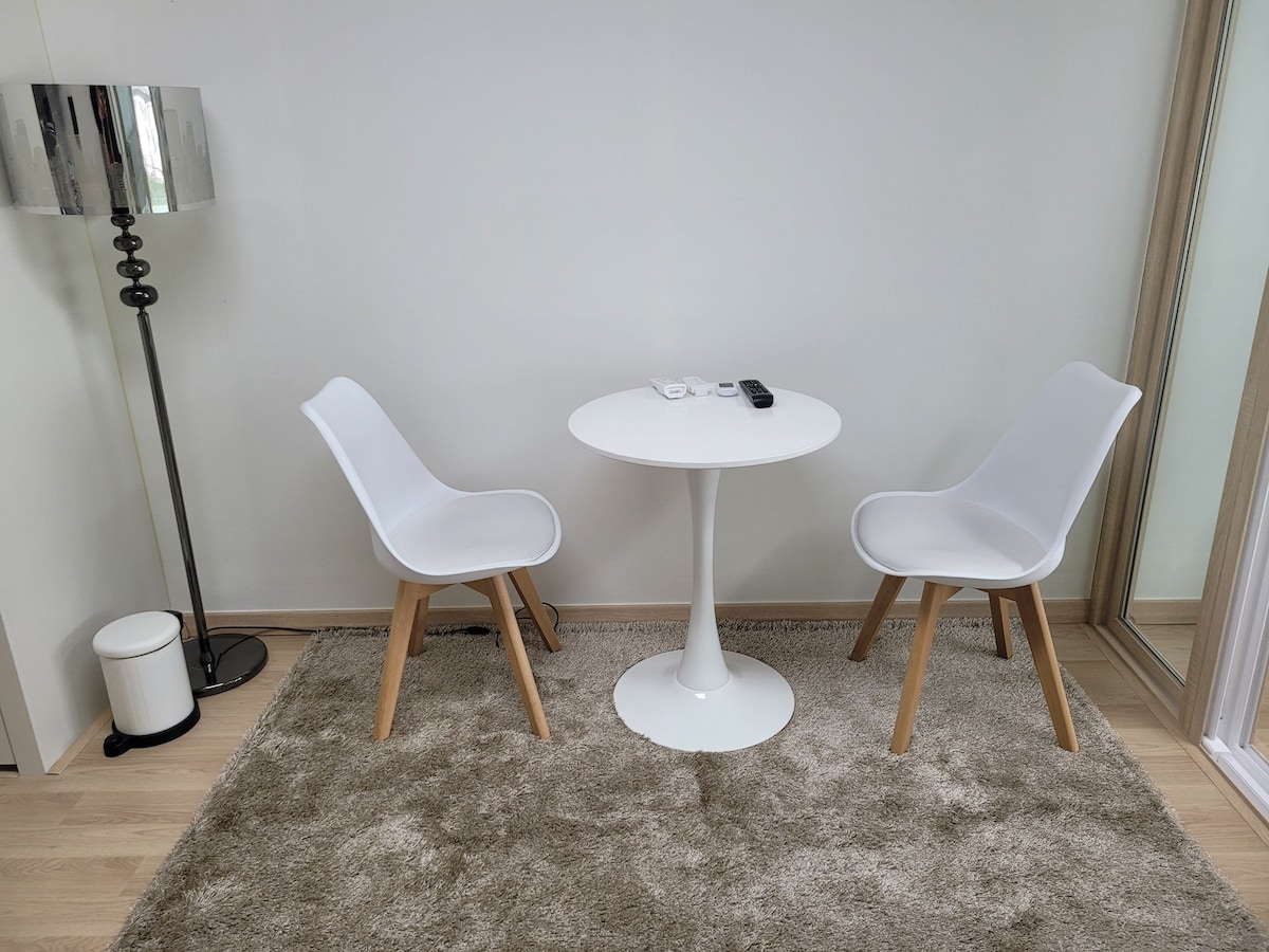 A simple seating area features two white chairs with wooden legs positioned around a round, white table. A floor lamp with a metallic shade stands beside the table, next to a small white trash can. The light-colored carpet adds a soft touch to the space.