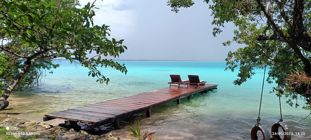 A wooden dock extends into crystal clear water, flanked by lush greenery. Two lounge chairs are positioned at the dock's end, providing a serene spot to enjoy the surrounding natural beauty.