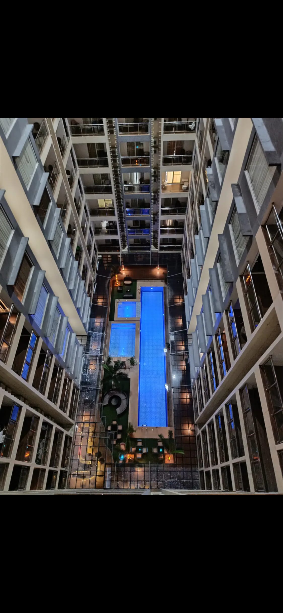 An aerial view captures the central courtyard of the building, featuring illuminated swimming pools surrounded by greenery. The area is enhanced by warm lighting from the surrounding balconies, creating a serene atmosphere in the evening. The layout reflects a well-planned outdoor space.